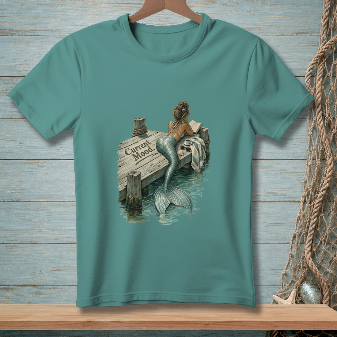 Teal t-shirt with a mermaid reading on a dock, text "Current Mood"