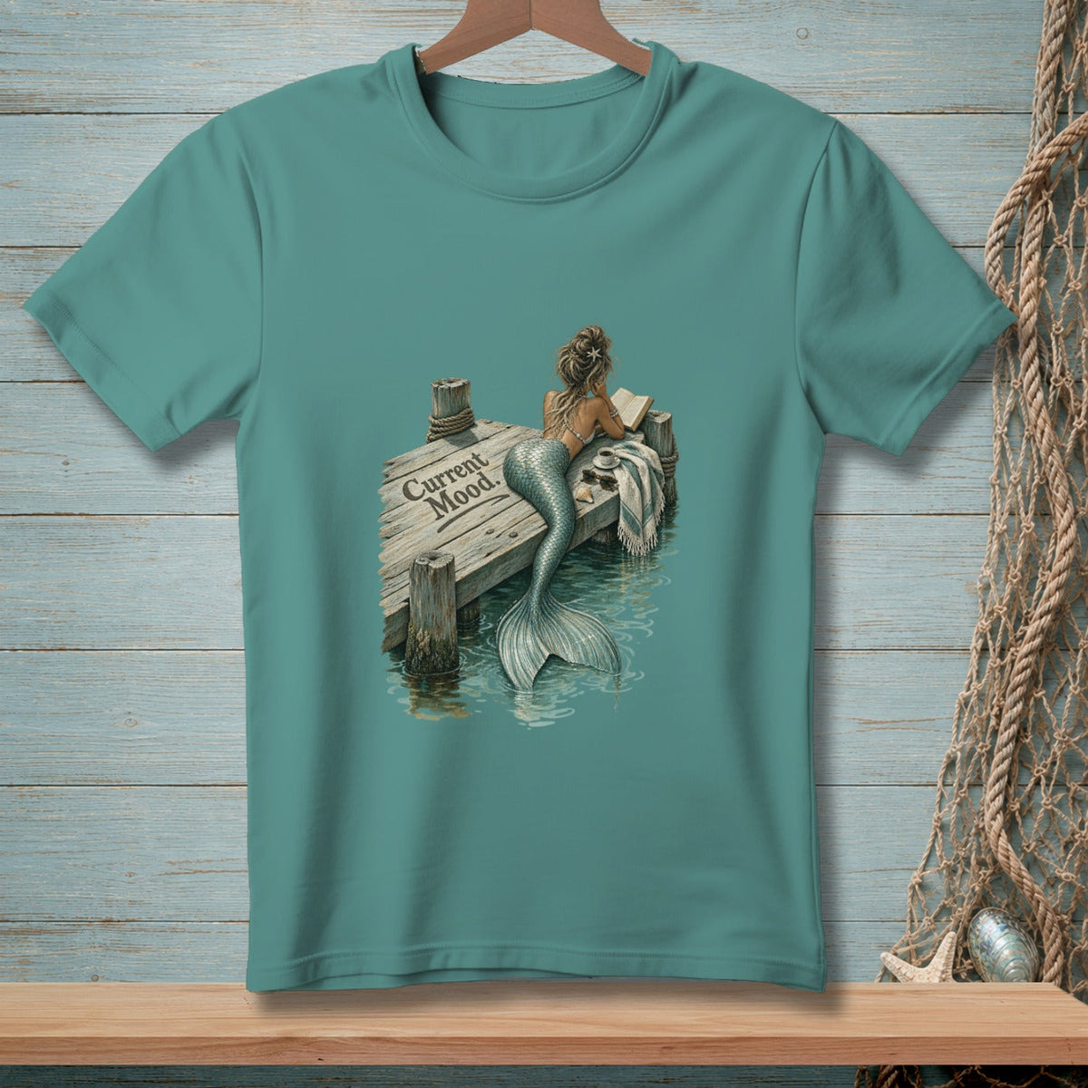 Teal t-shirt with a mermaid reading on a dock, text "Current Mood"