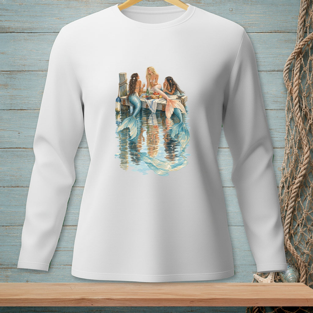 Three mermaids sit on a dock, with reflections in the water.