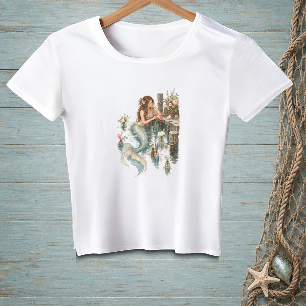 White t-shirt with a mermaid sitting by a dock surrounded by flowers.
