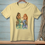 Two mermaids with colorful tails sit on a wooden dock, sharing a bowl of fruit.
