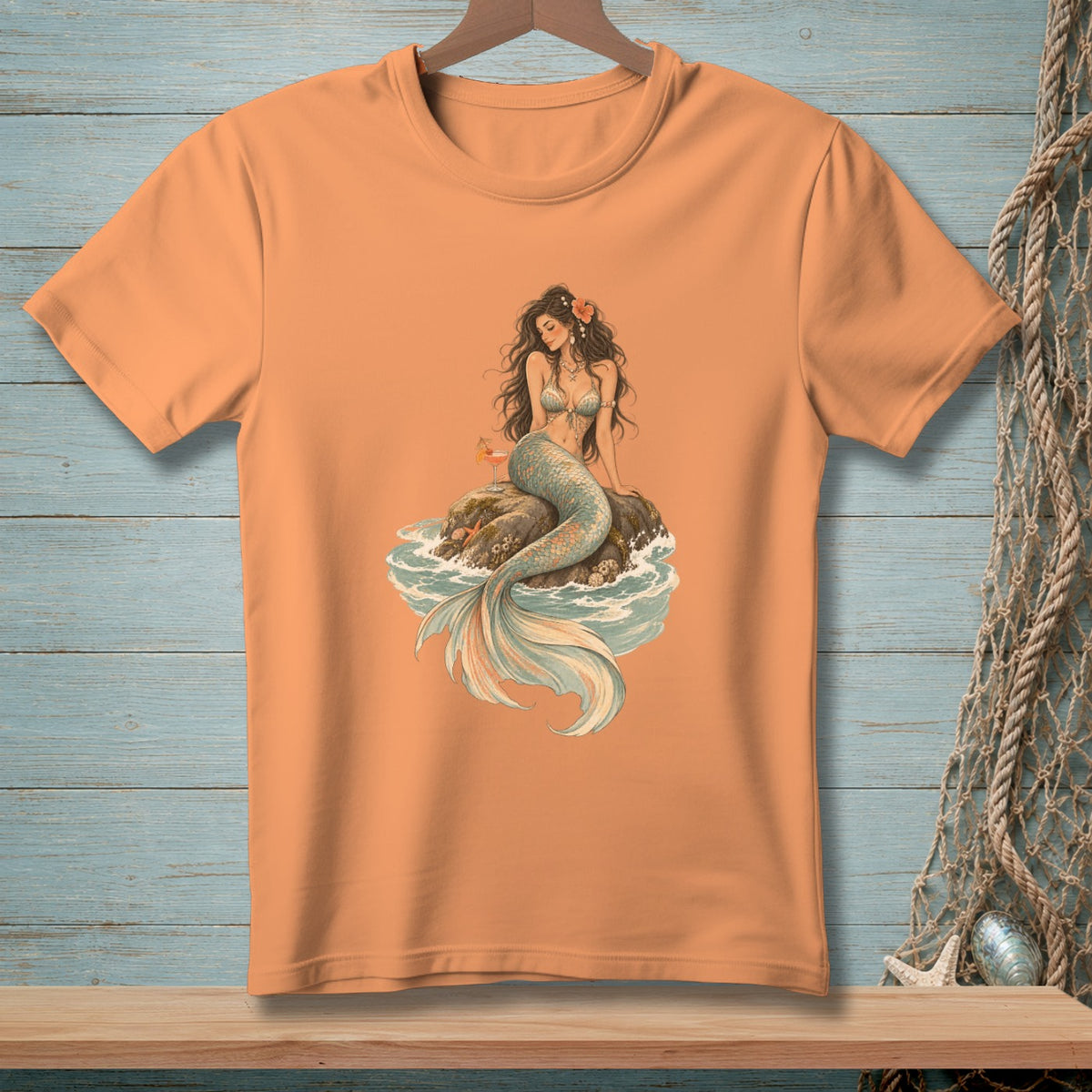 Peach t-shirt with a drawing of a mermaid sitting on a rock by the sea.