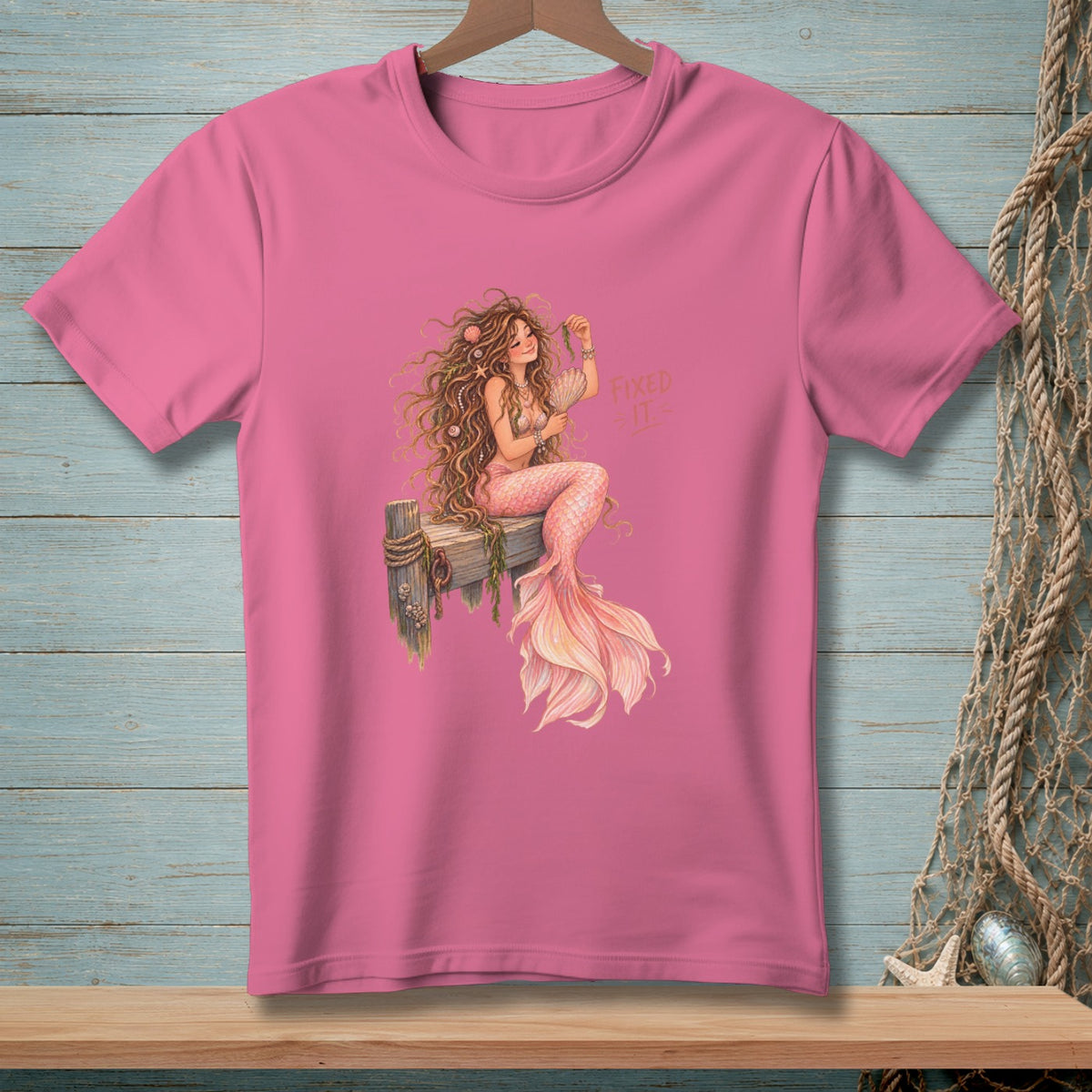 Pink t-shirt with illustration of a mermaid sitting on a wooden post.