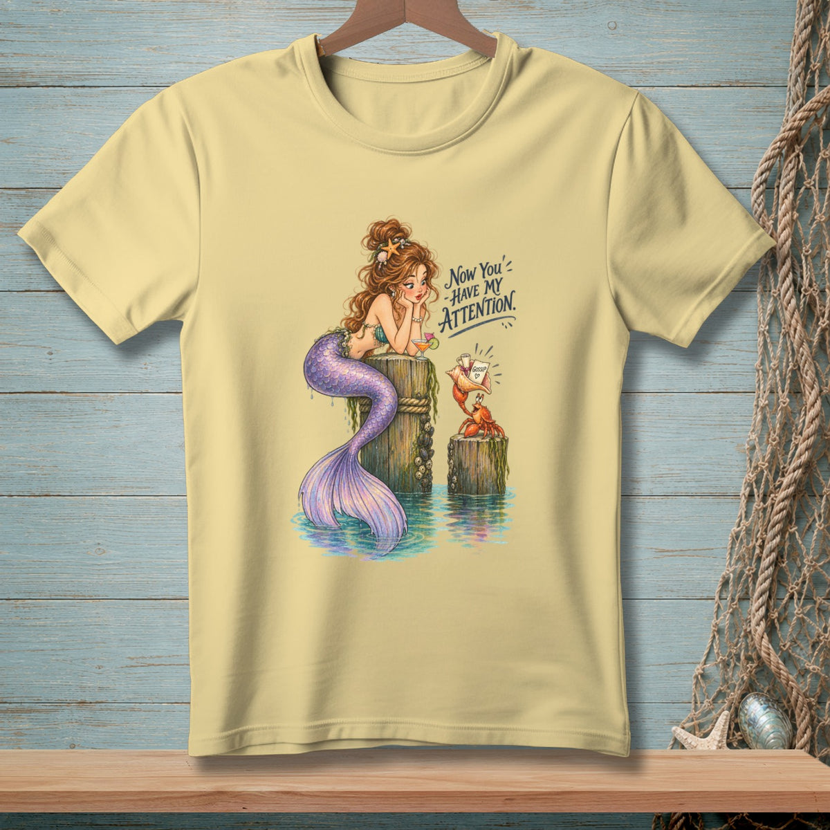 Light yellow t-shirt with mermaid and crab illustration, text "Now You' HAVE MY ATTENTION."