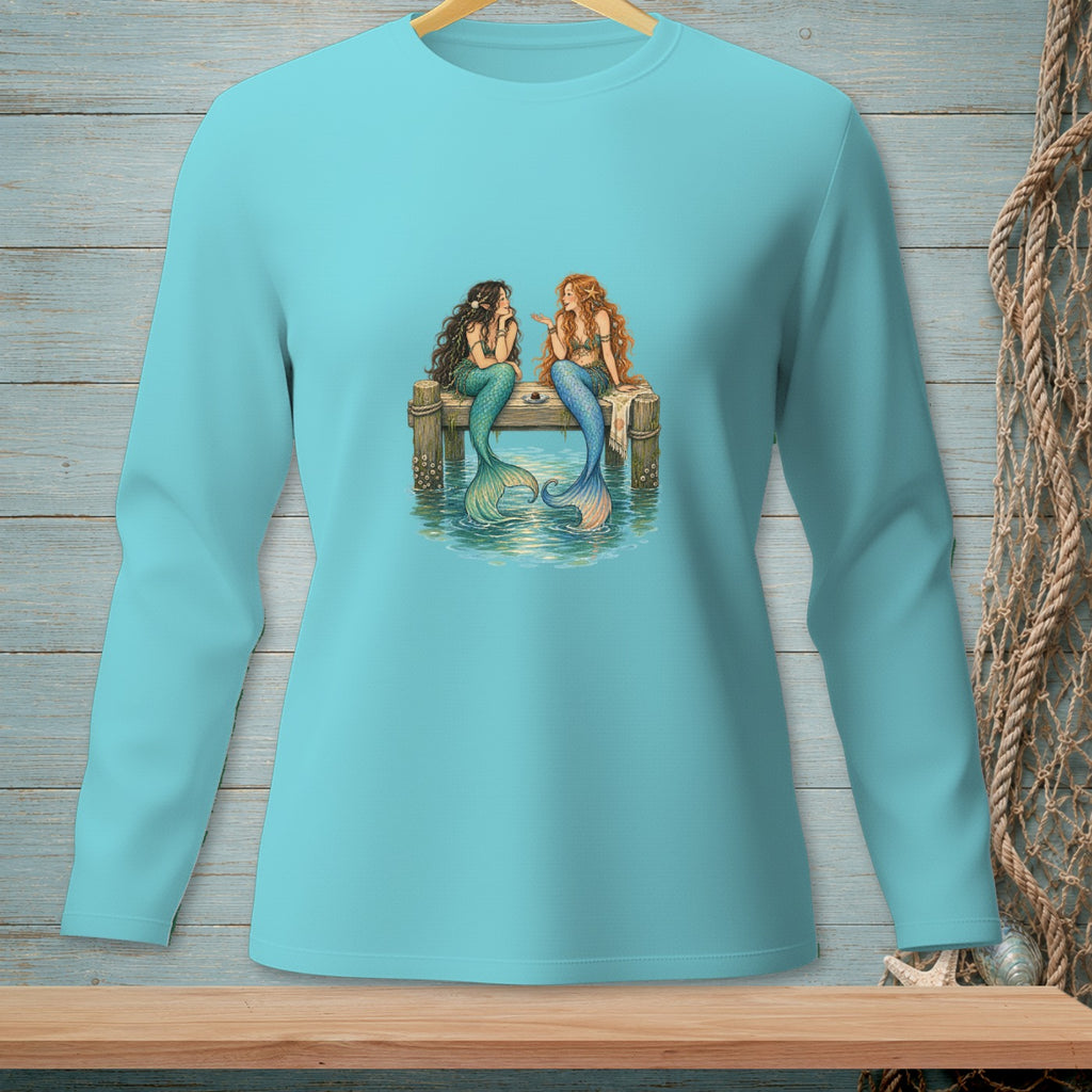 Two mermaids with tails sit talking on a wooden dock in ocean water.