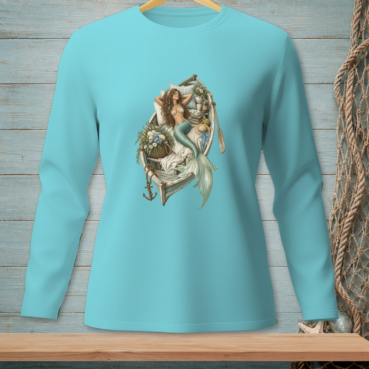 Mermaid lounging in a boat with flowers, oars, and anchor graphic on a light blue long-sleeve shirt.