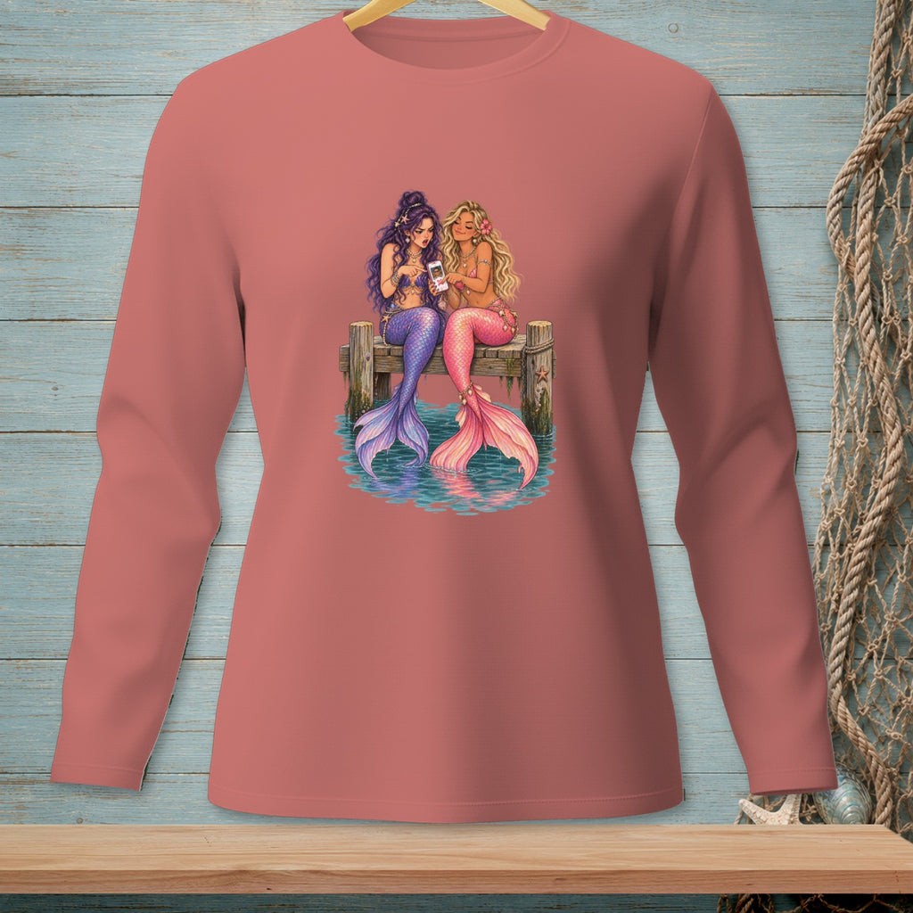 Two mermaids with purple and pink tails sit on a dock looking at a phone.