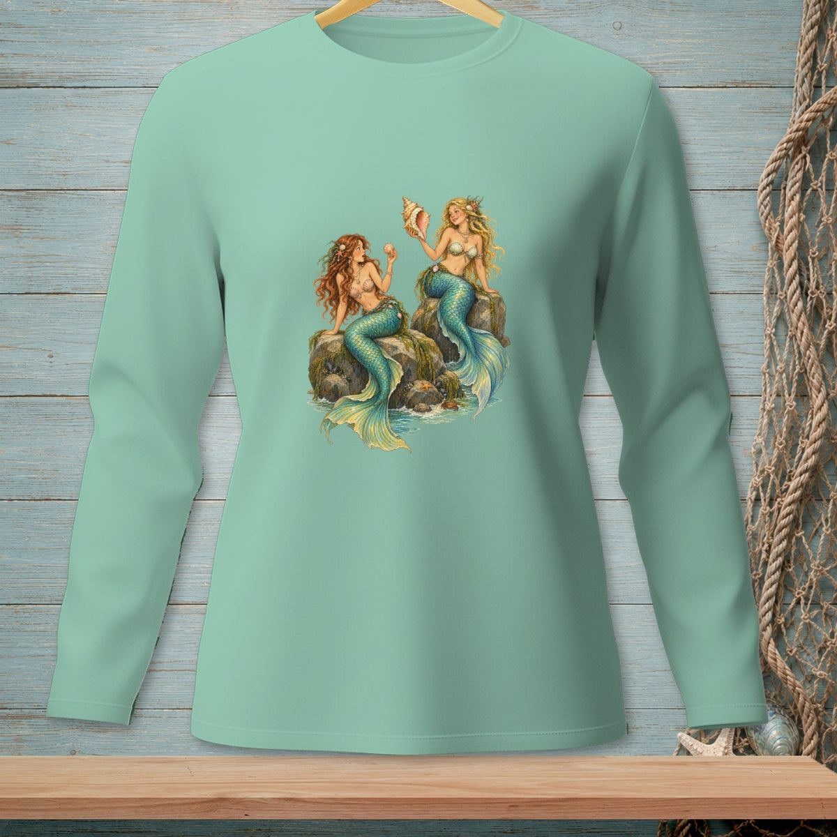 Two mermaids sitting on rocks, one holding a seashell, on a light green long-sleeved shirt.