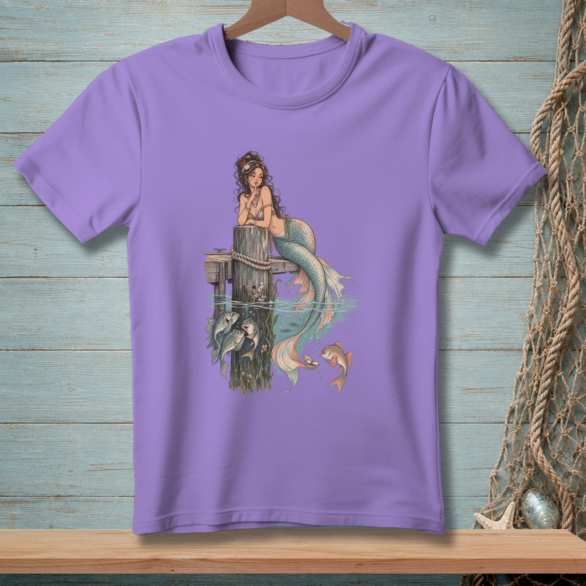 Purple t-shirt with a mermaid illustration resting on a wooden post by the water.