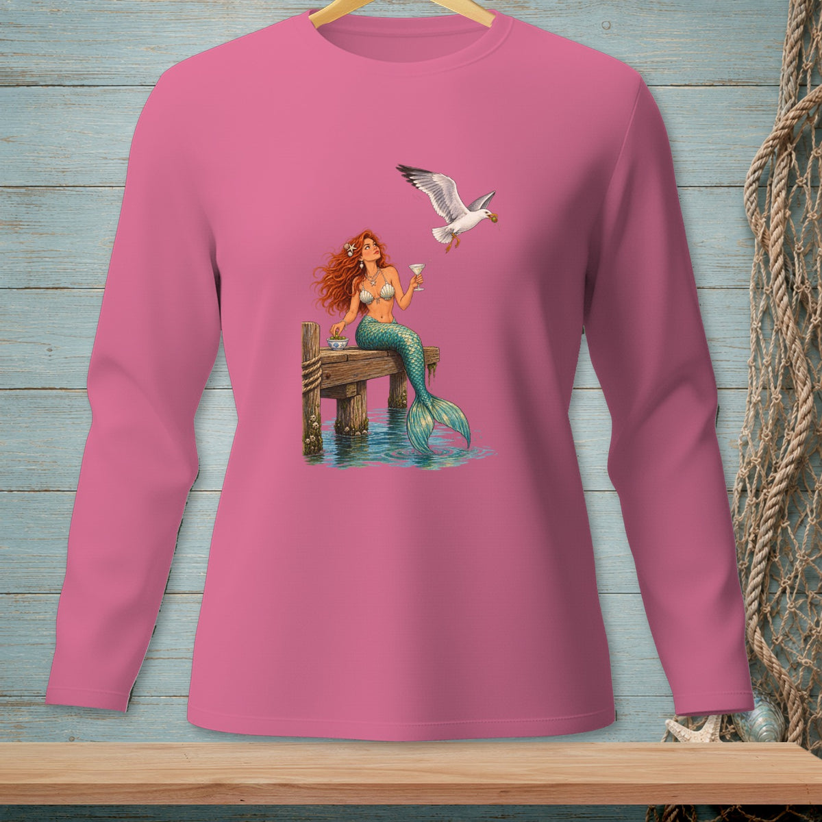 Mermaid with long red hair sits on dock with drink and food as seagull flies by.