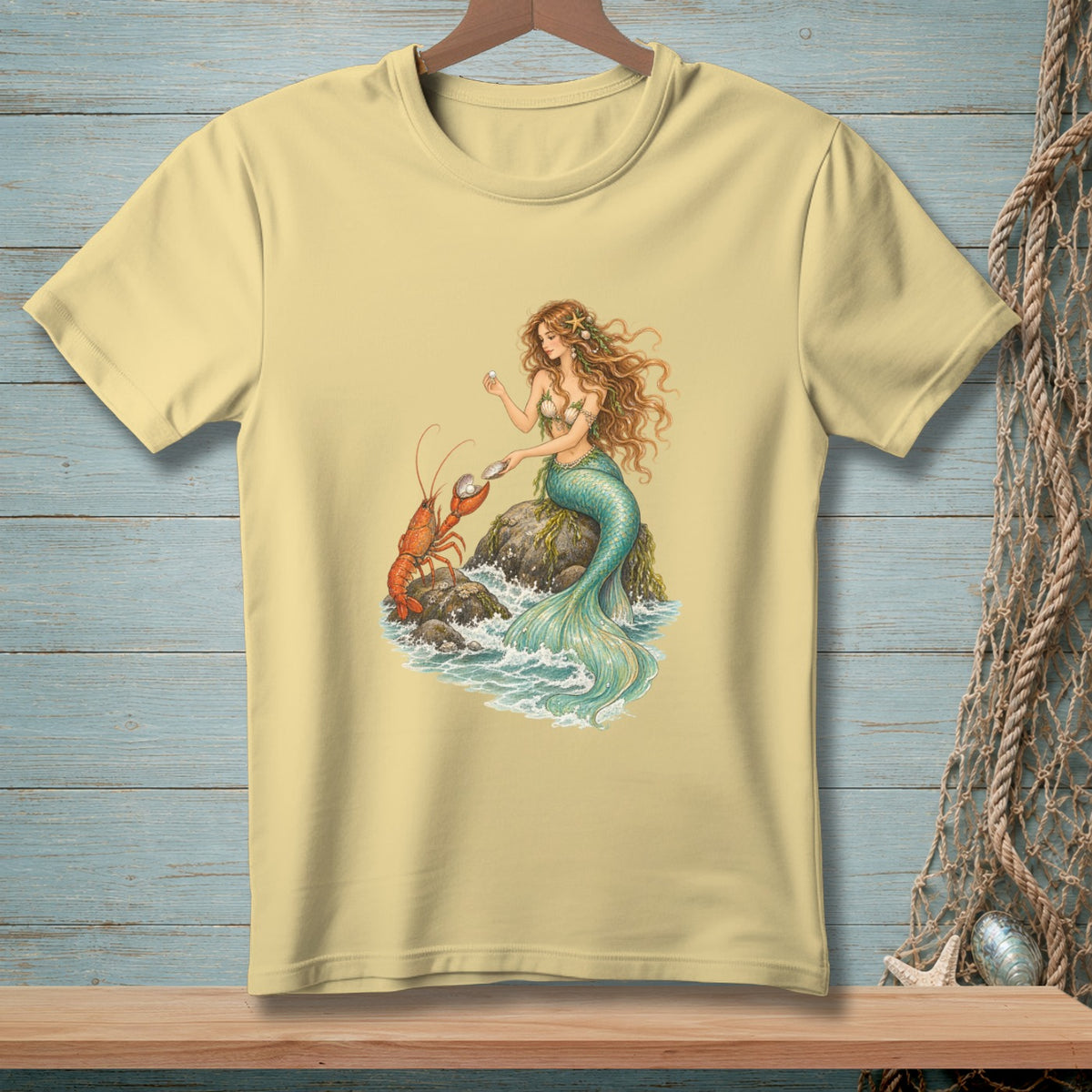 Cream t-shirt with mermaid and lobster illustration on a rock by the sea.