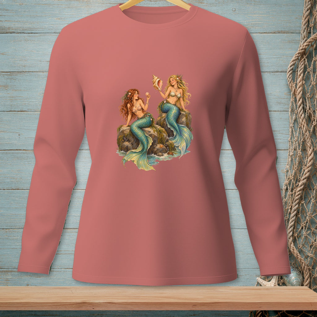 Two mermaids sit on rocks, one holding a seashell, on a pink long-sleeved shirt.