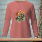 Two mermaids sit on rocks, one holding a seashell, on a pink long-sleeved shirt.
