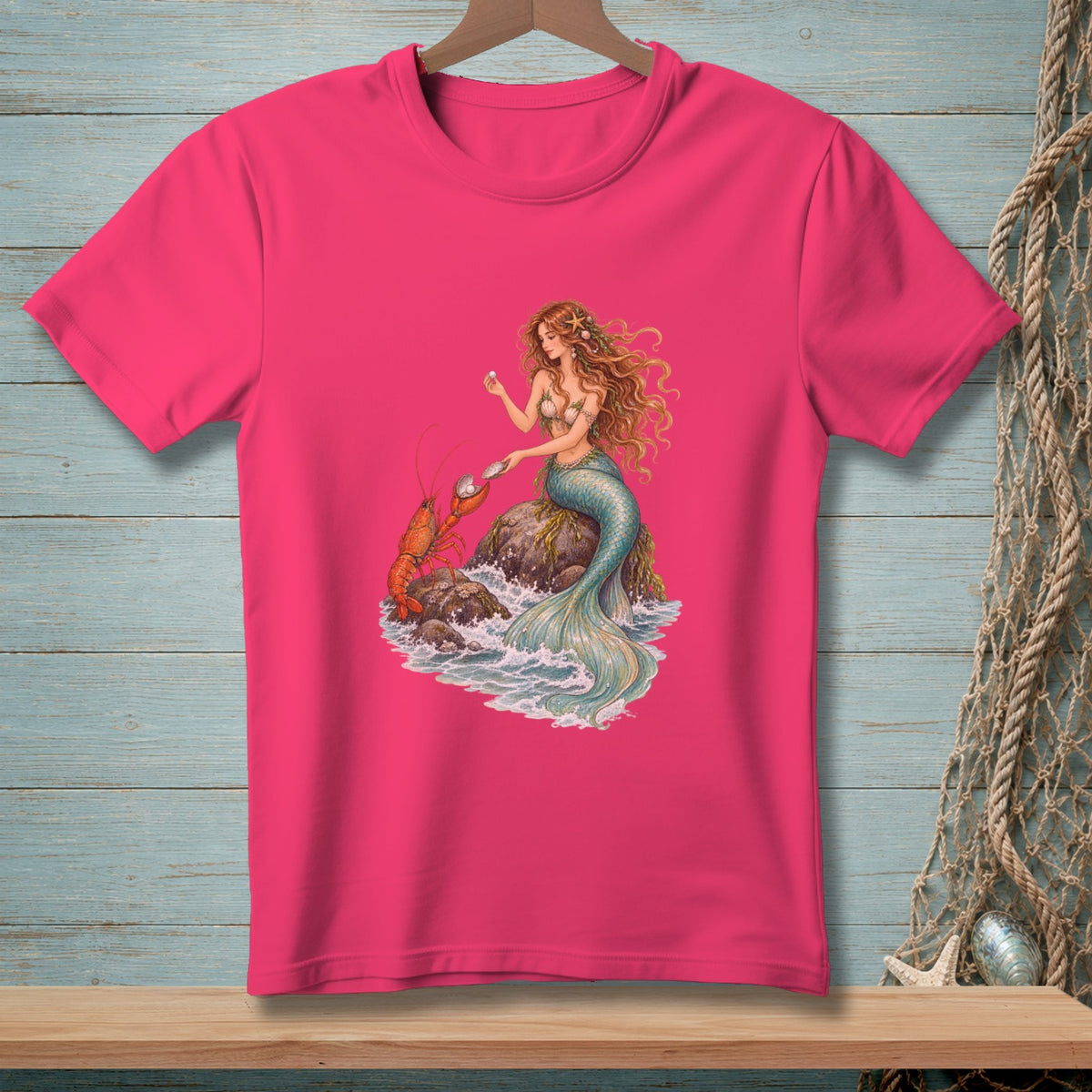 Pink t-shirt with a mermaid and lobster illustration on rocks with waves.