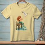 Pale yellow t-shirt with a mermaid sitting on a dock while a seagull flies overhead.