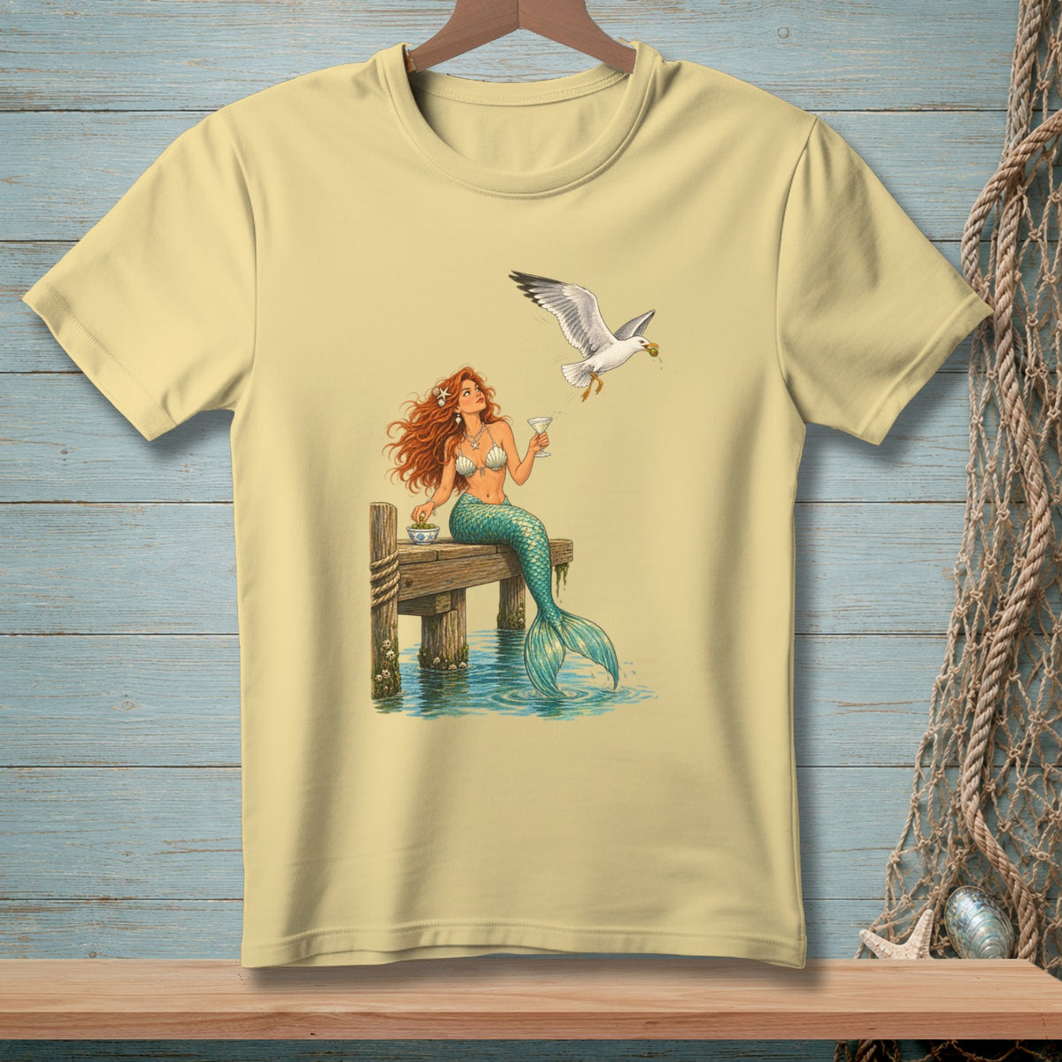 Pale yellow t-shirt with a mermaid sitting on a dock while a seagull flies overhead.