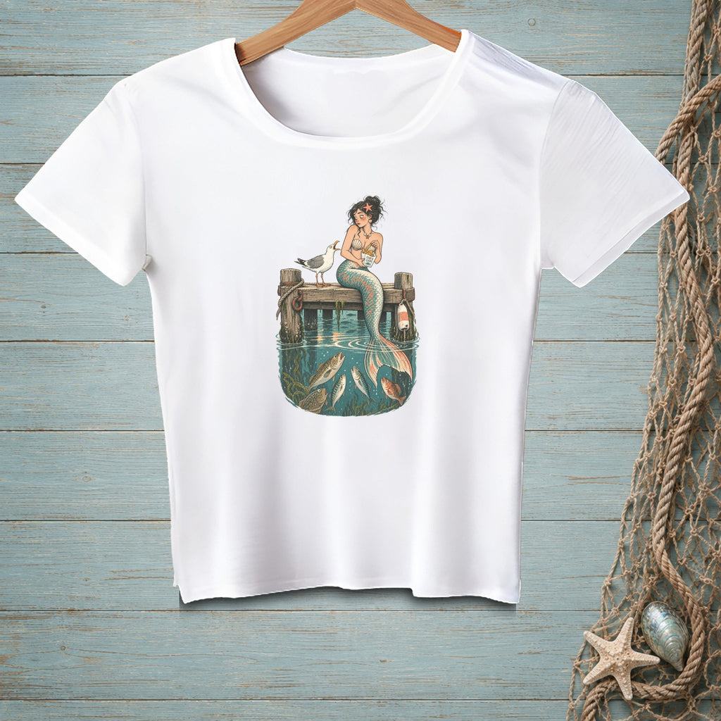 Mermaid sitting on dock feeding seagull, with fish swimming in the water below.