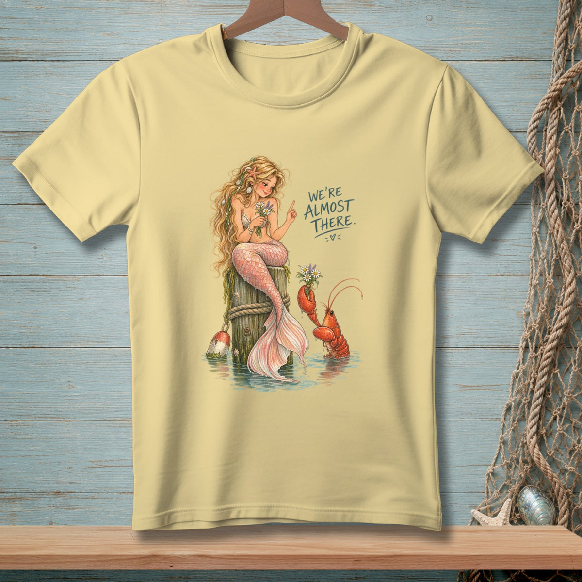 Light yellow t-shirt with mermaid and lobster graphic, text "WE'RE ALMOST THERE."