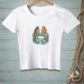 White t-shirt with two mermaids sitting on a dock talking.