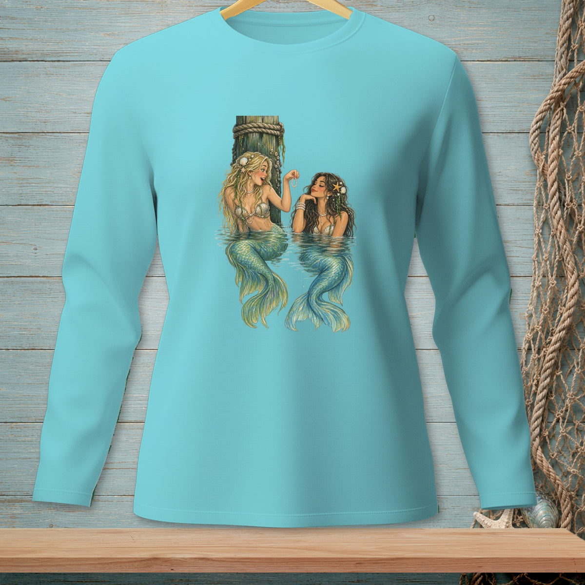 Two mermaids with colorful tails sit in water by a wooden post on a light blue shirt.