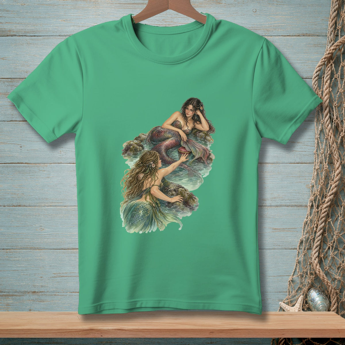 Green t-shirt with an illustration of two mermaids on rocks in water.