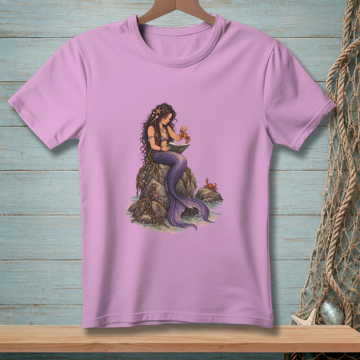 Mermaid sitting on rocks reading book with crabs nearby on pink t-shirt.