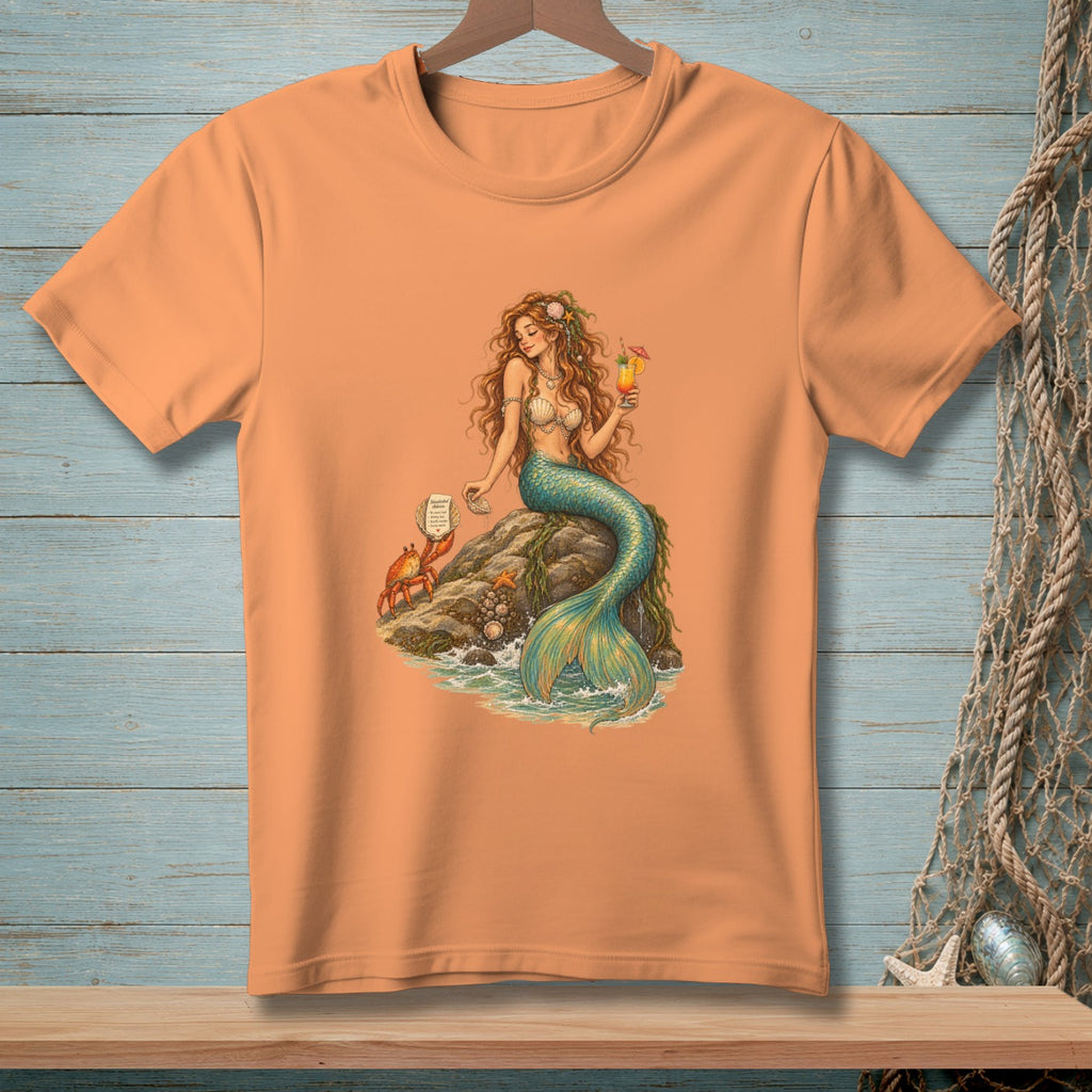 Peach t-shirt with mermaid, crab, and cocktail graphic on a rock.