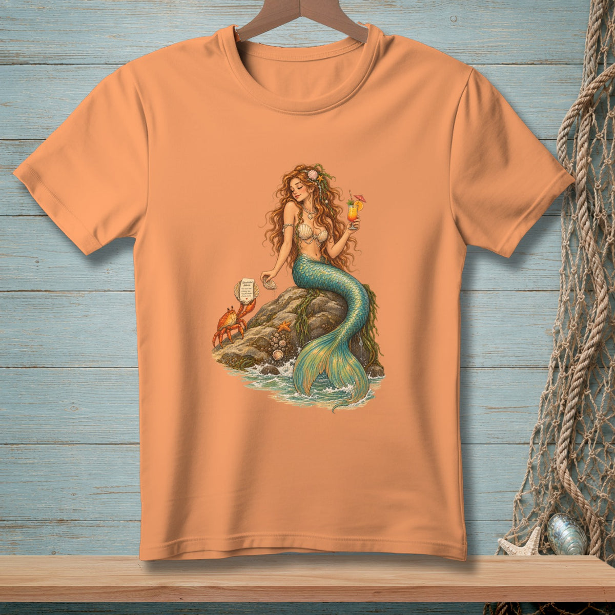 Peach t-shirt with mermaid, crab, and cocktail graphic on a rock.