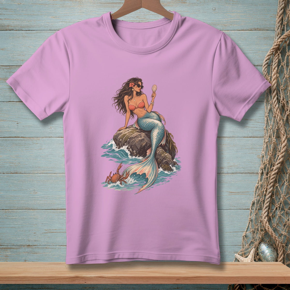 Purple t-shirt with a mermaid holding a seashell on a rock, with a crab in the water.