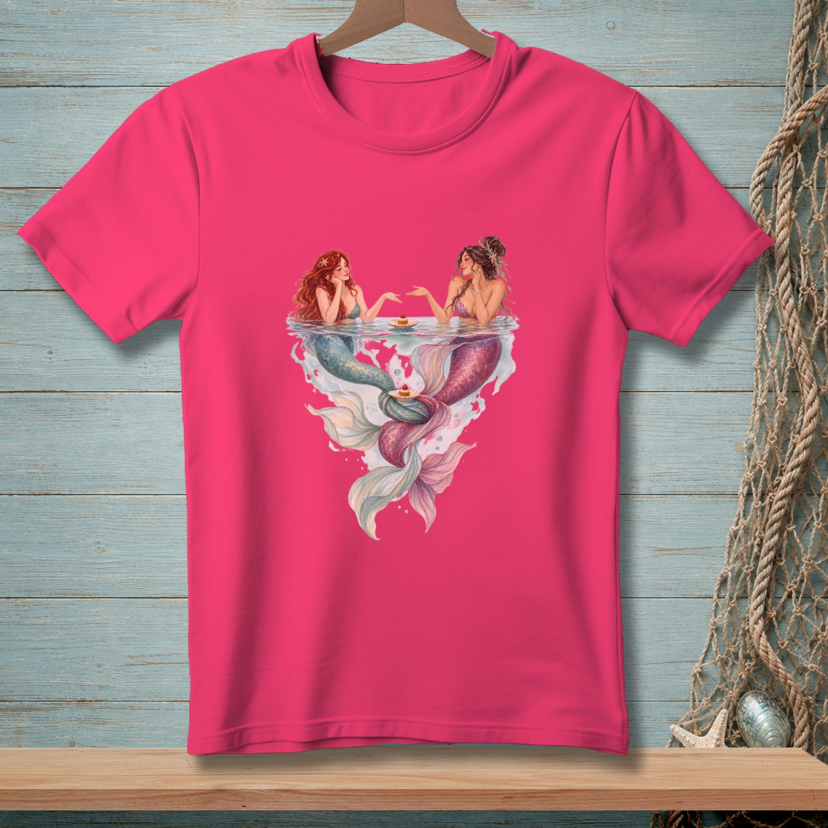 Two mermaids with cakes float in heart-shaped water on a pink t-shirt.