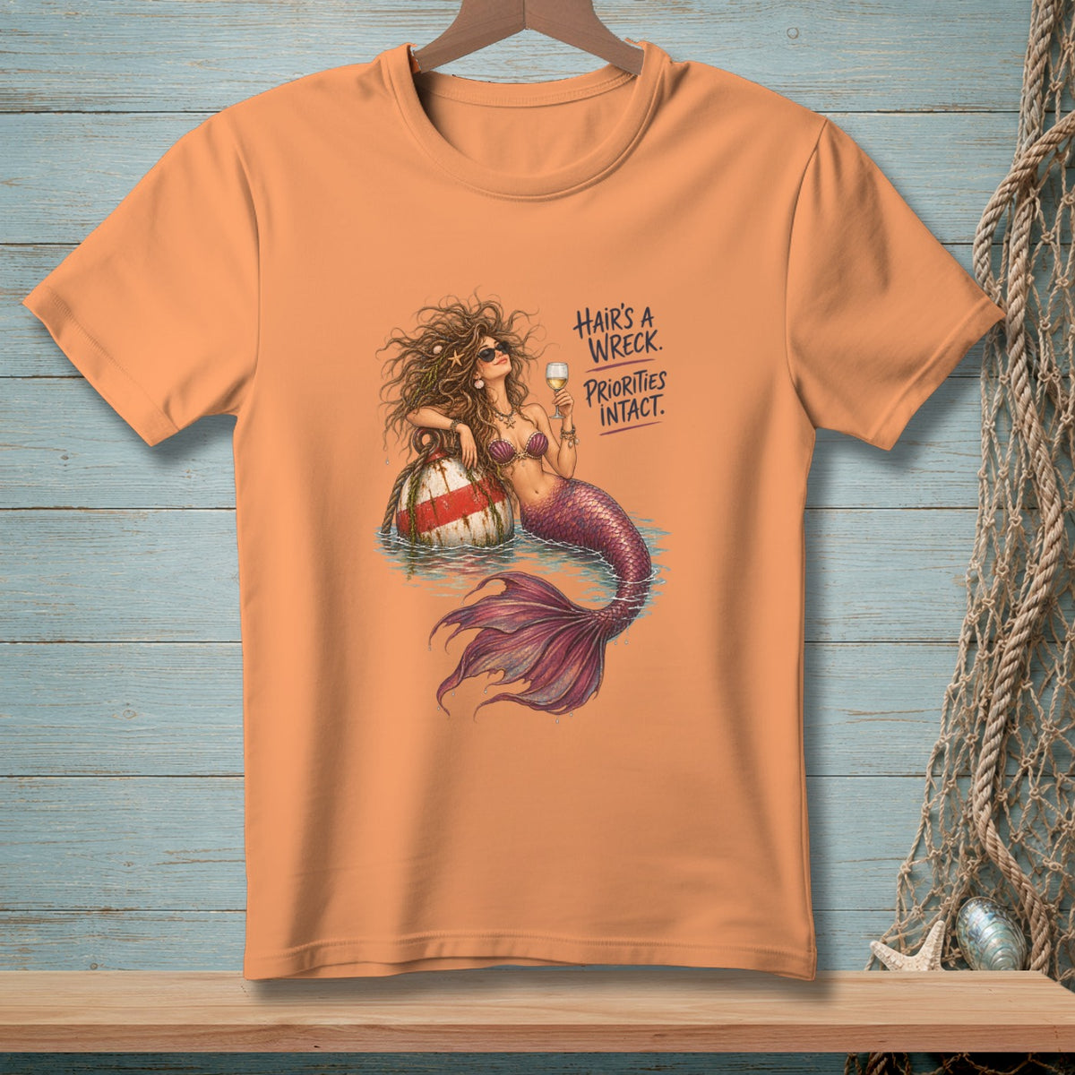 Peach t-shirt with a mermaid on a buoy and text: Hair's a wreck. Priorities intact.