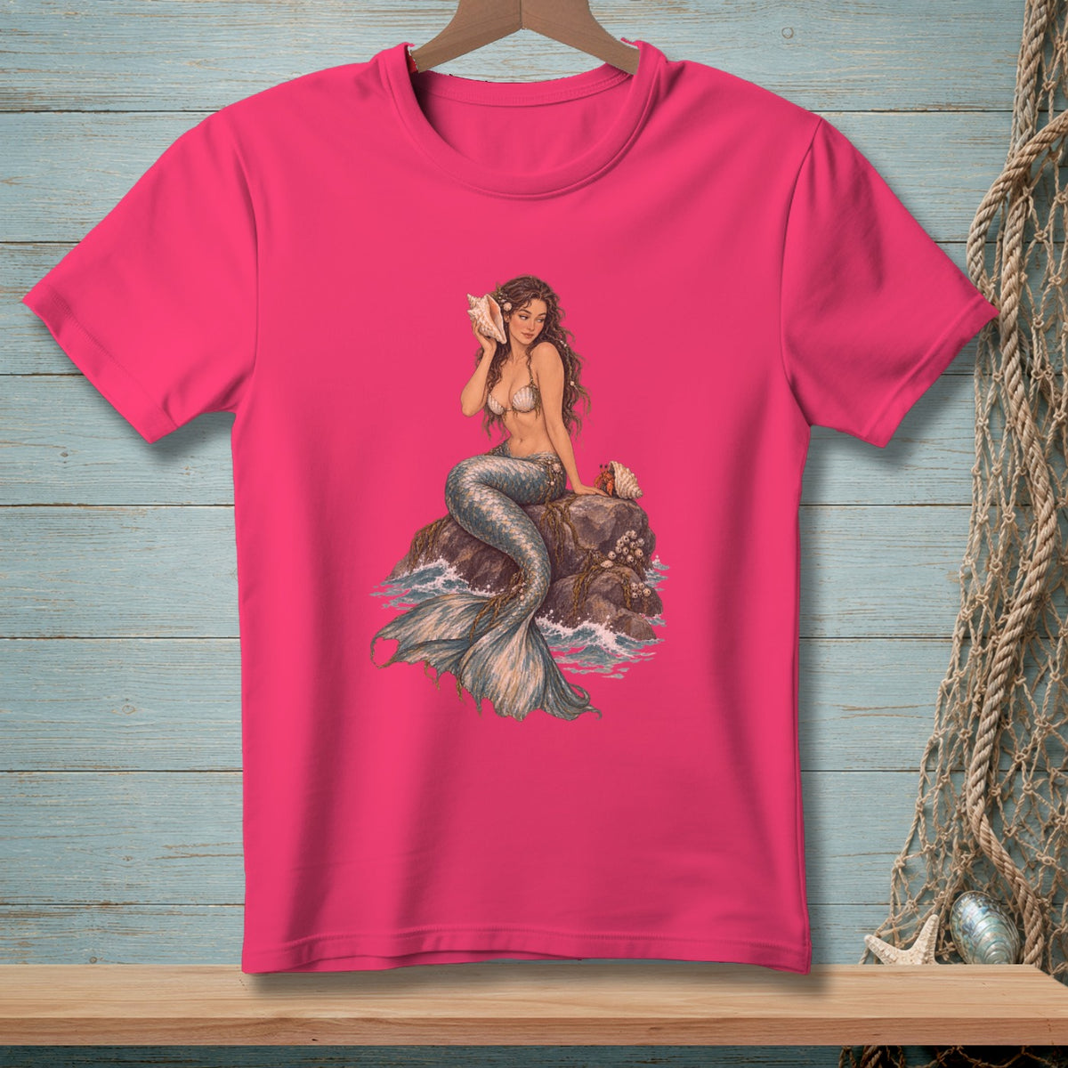 Pink t-shirt with a graphic of a mermaid sitting on a rock holding a seashell.