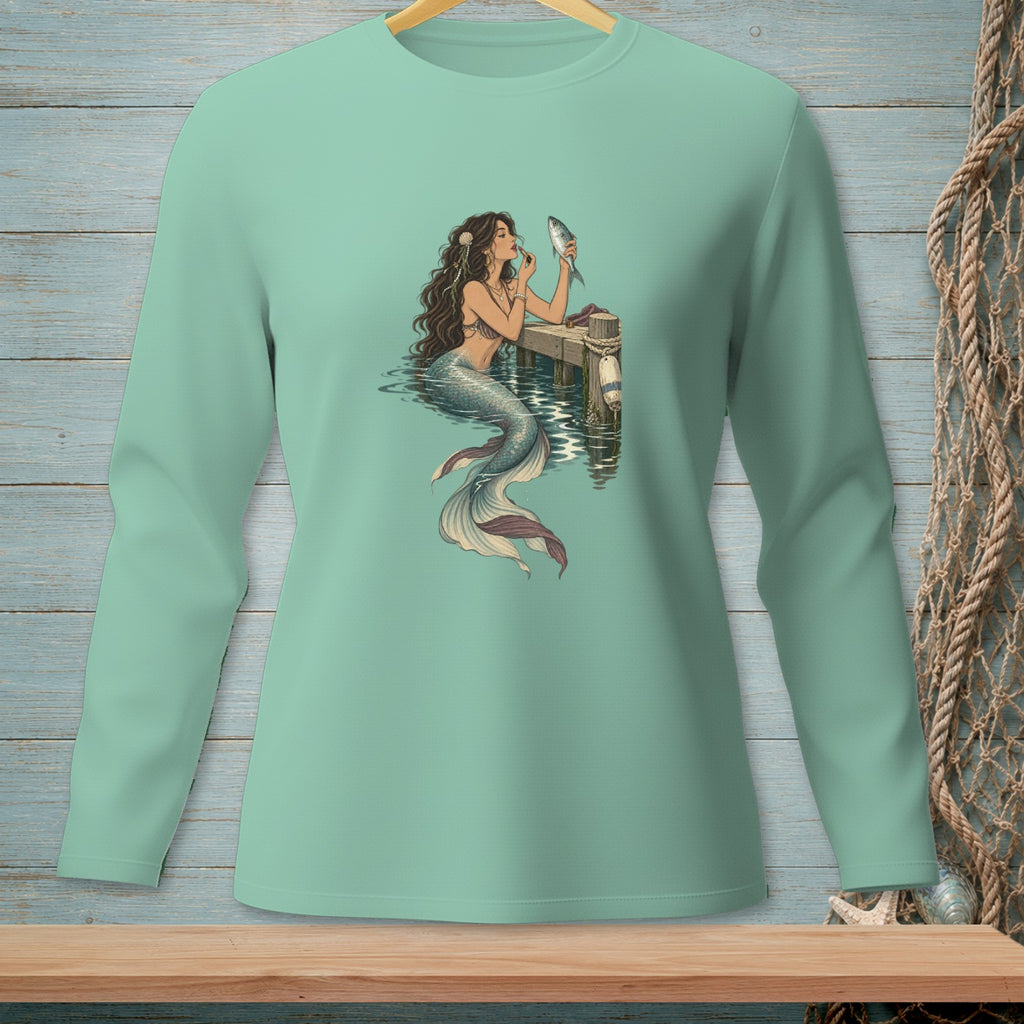Mermaid applying makeup on a dock with a fish, on a light teal long-sleeve shirt.