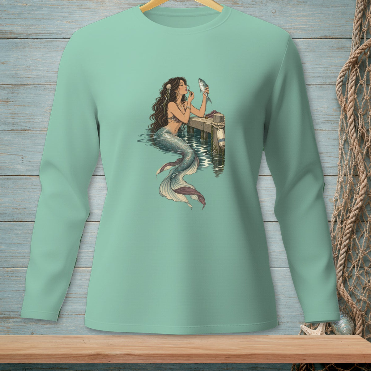 Mermaid applying makeup on a dock with a fish, on a light teal long-sleeve shirt.