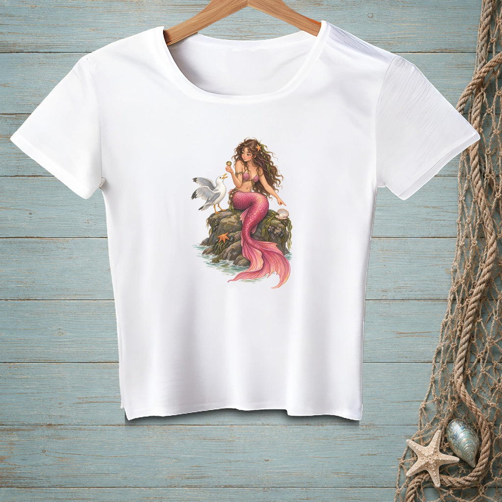 White t-shirt with a printed illustration of a mermaid sitting on rocks with a seagull and a seashell.