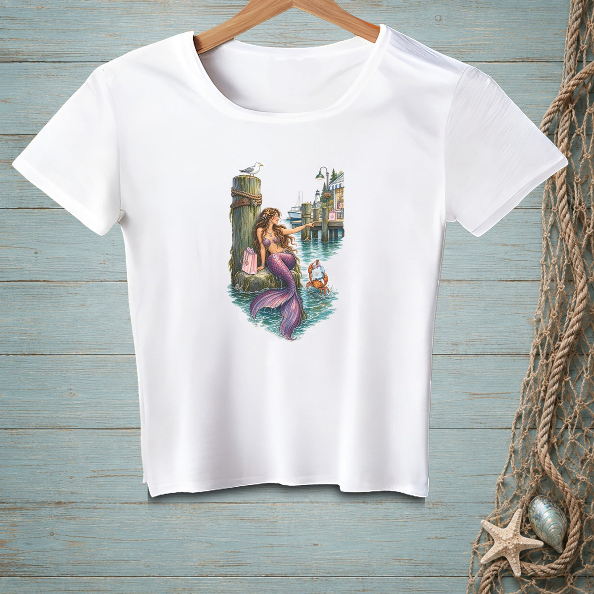 White t-shirt featuring a mermaid with a purple tail, sitting on a dock with a shopping bag.