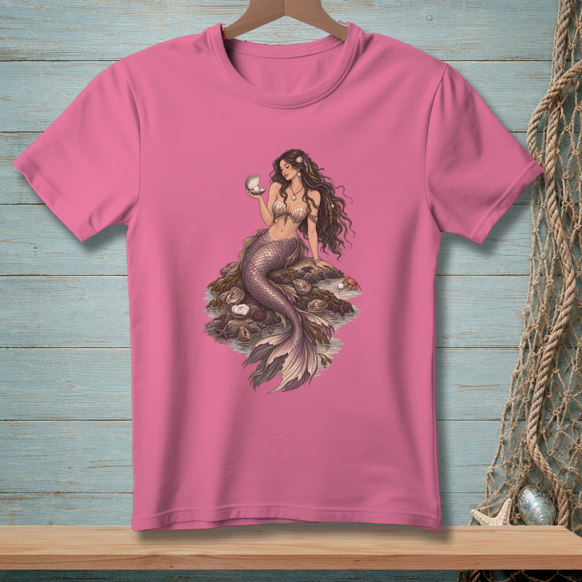 Pink t-shirt with mermaid holding a pearl sitting on rocks and shells.
