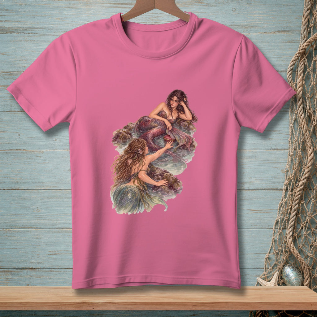 Pink t-shirt with an illustration of two mermaids interacting on rocks by the water.