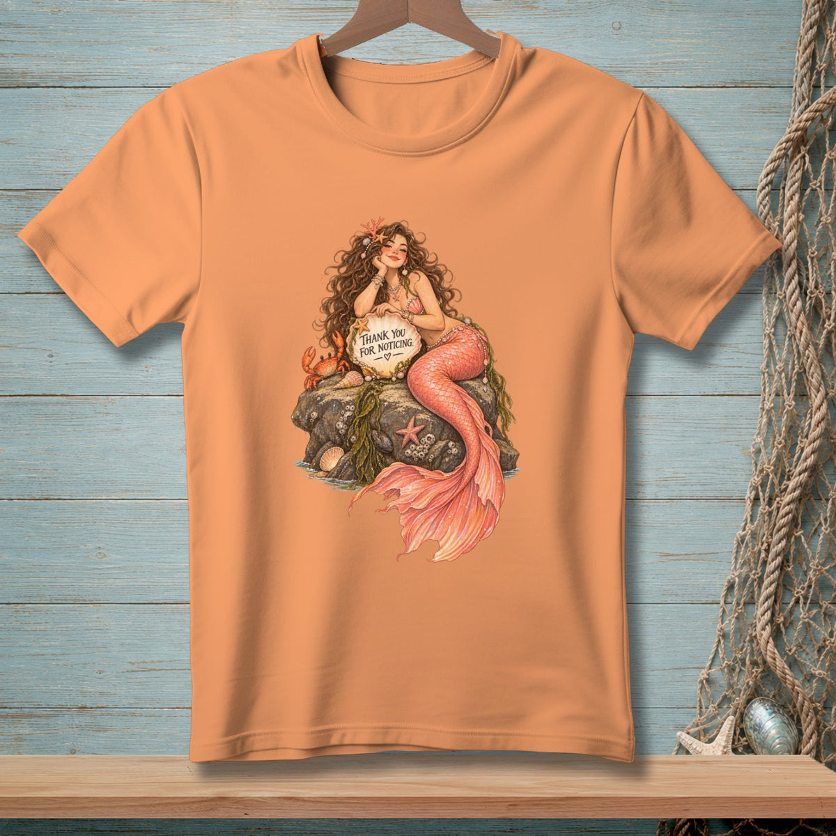 Peach t-shirt with illustration of a mermaid sitting on rocks with a crab, holding a sign.