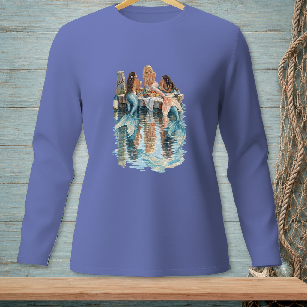 Three mermaids with colorful tails sit at a table on a dock with water reflections.