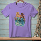 Two mermaids with teal tails sit on a dock, drinking wine by the water.