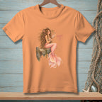 Peach t-shirt with a watercolor illustration of a mermaid sitting on a wooden dock.