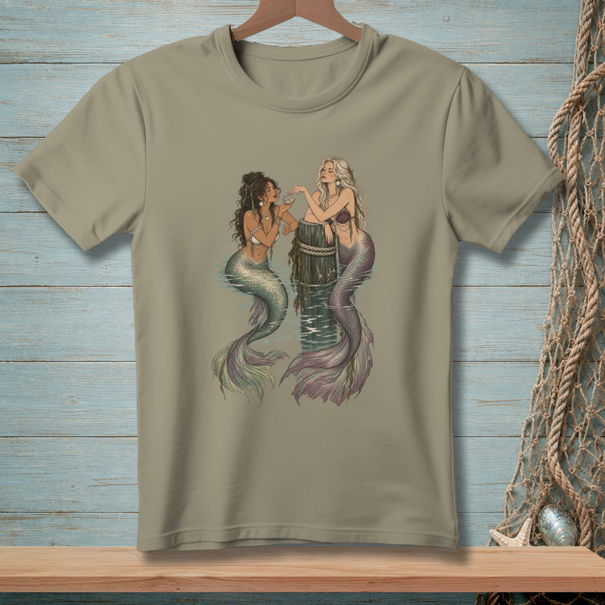 T-shirt graphic features two mermaids sitting by a wooden post in water.