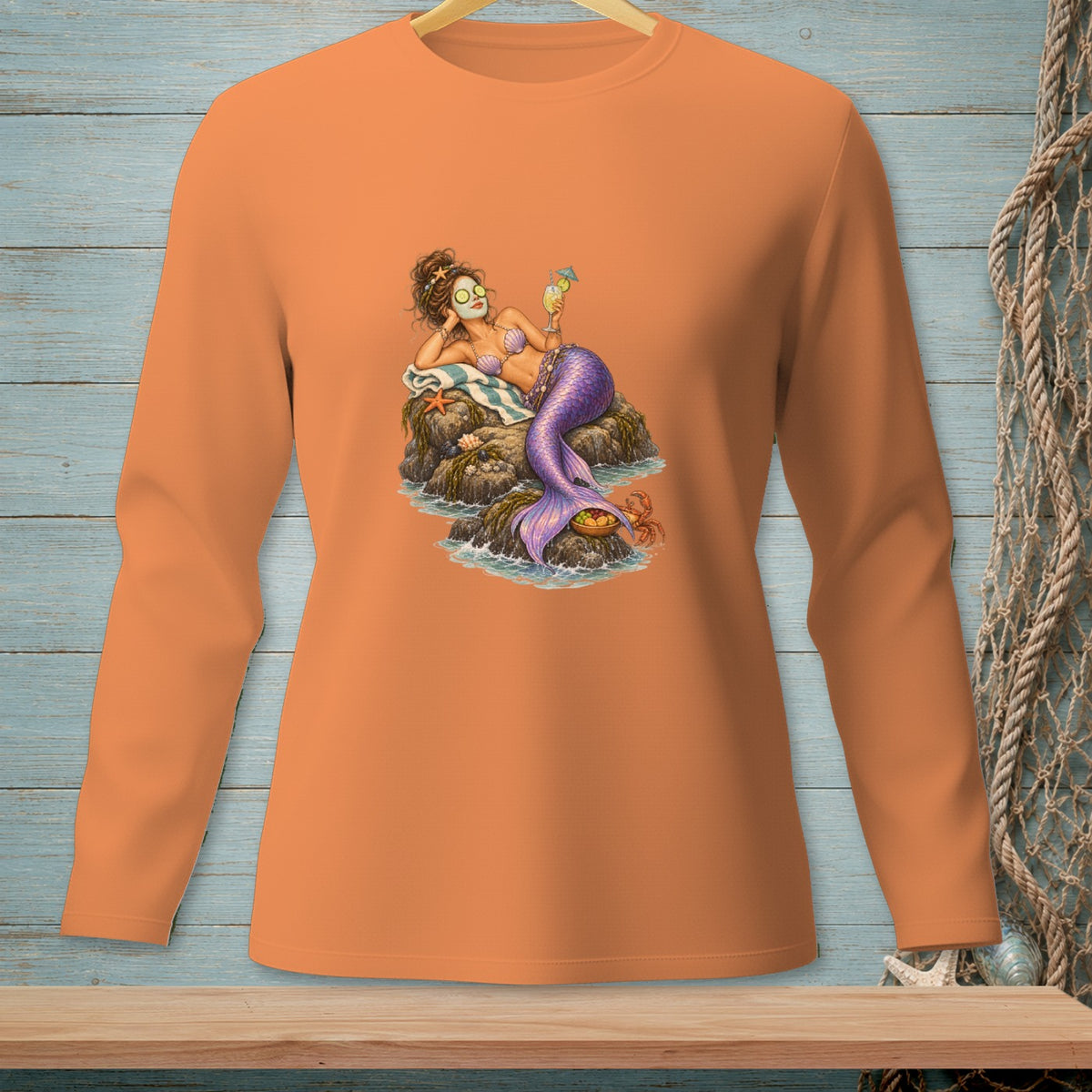 Orange long-sleeve shirt featuring a mermaid relaxing on rocks with a drink and face mask.