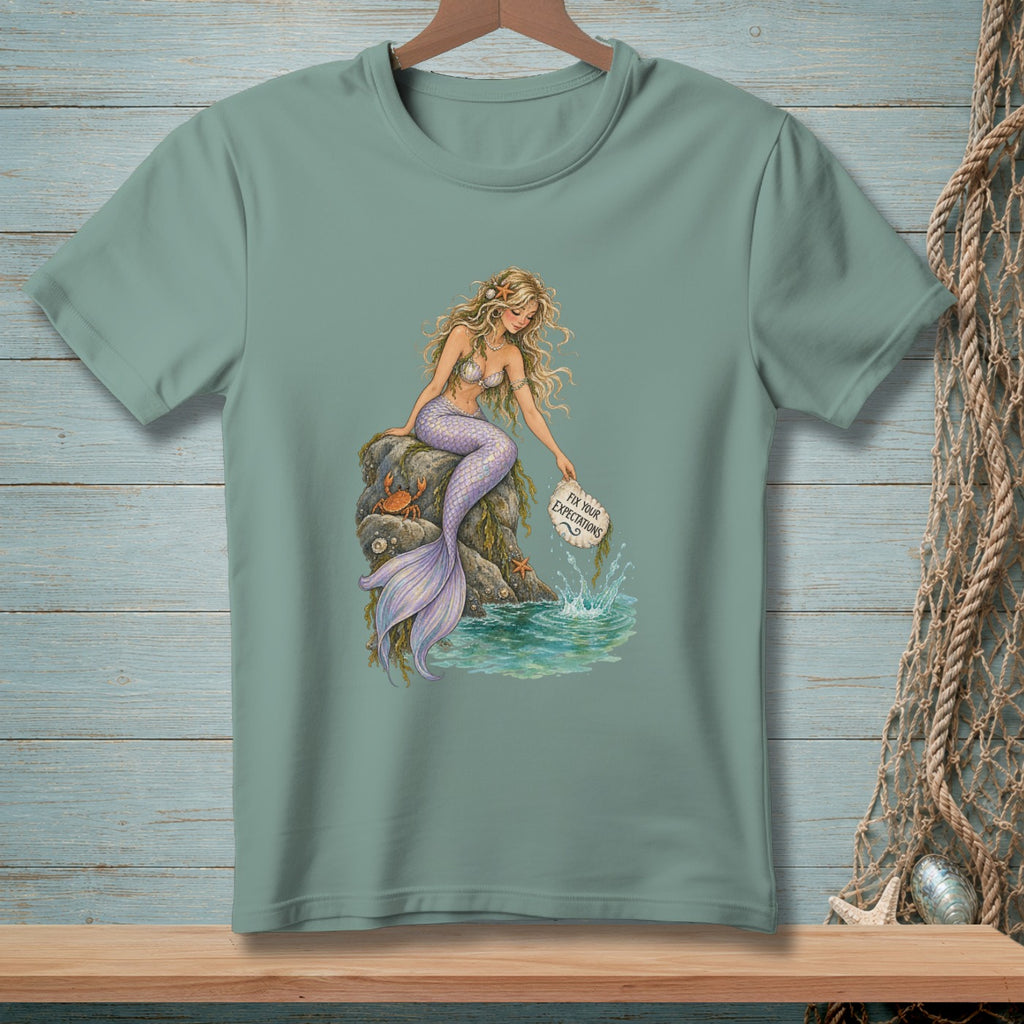 Mermaid sitting on rocks by the ocean holding a sign that says "Fix Your Expectations".