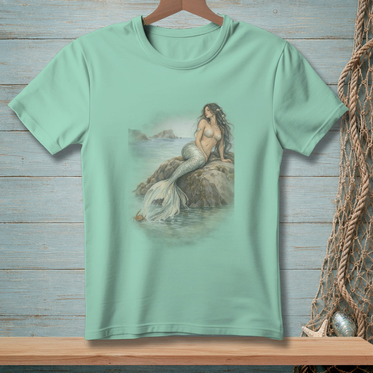 Mint green t-shirt with a mermaid sitting on a rock by the sea.