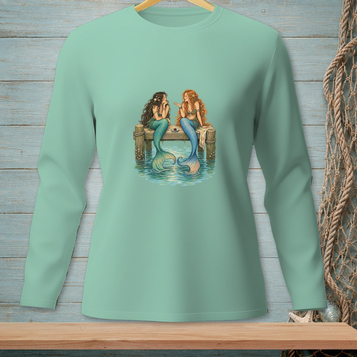 Two mermaids with tails sit on a dock, talking near a small table.