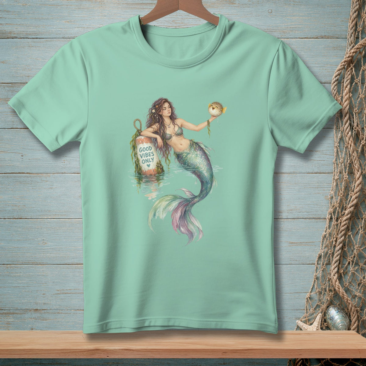Mint t-shirt with a watercolor mermaid holding a pufferfish near a buoy that reads "GOOD VIBES ONLY."