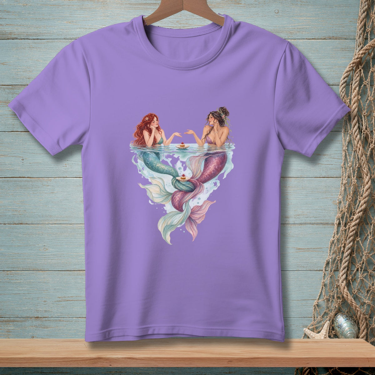 Lavender t-shirt with illustration of two mermaids holding cupcakes in water.