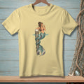 Light yellow t-shirt with a mermaid illustration holding a drink, leaning on a wooden post.